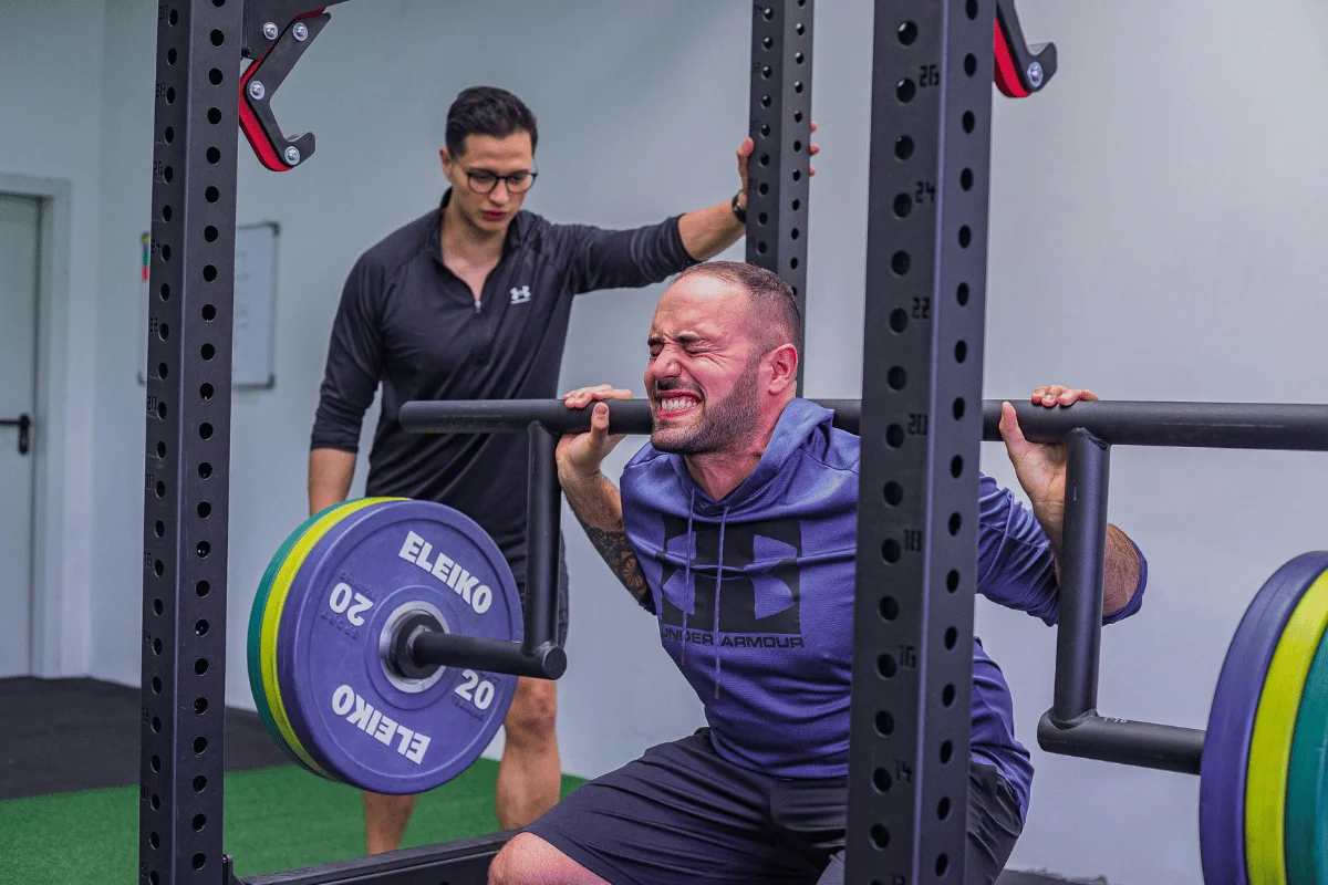 Man lifting weights on a squat rack with intense focus, while a coach monitors the session.