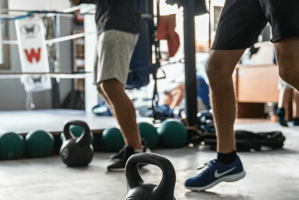 Close-up of two individuals exercising with kettlebells in a gym near a boxing ring and other fitness equipment.