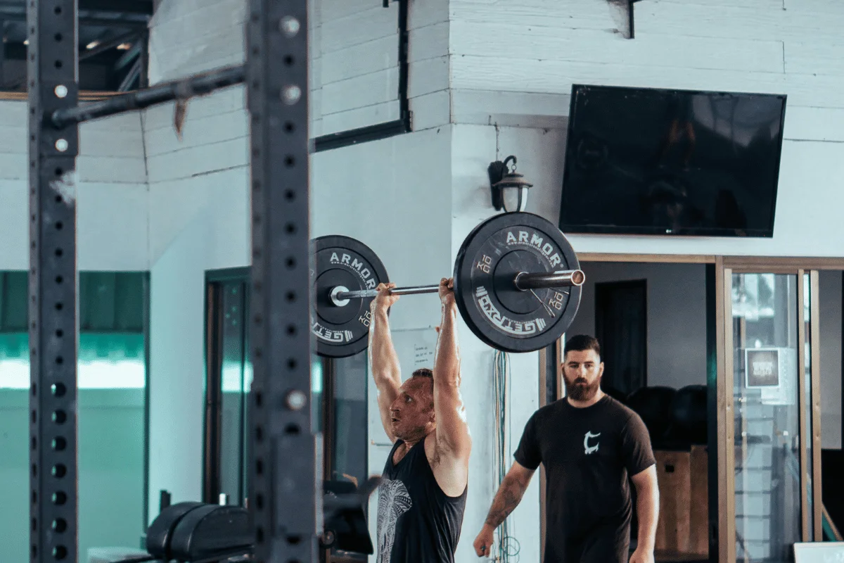 Man lifting a heavy barbell overhead while a trainer observes in a gym, performing a strength training exercise.