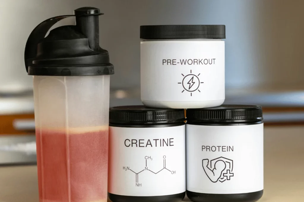 Pre-workout, creatine, and protein supplement containers next to a shaker bottle with a red drink.