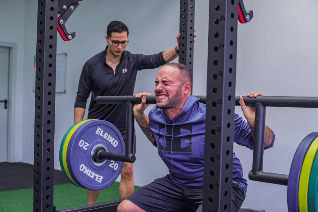 Man performing a barbell squat with heavy weights in a gym under the supervision of a trainer.