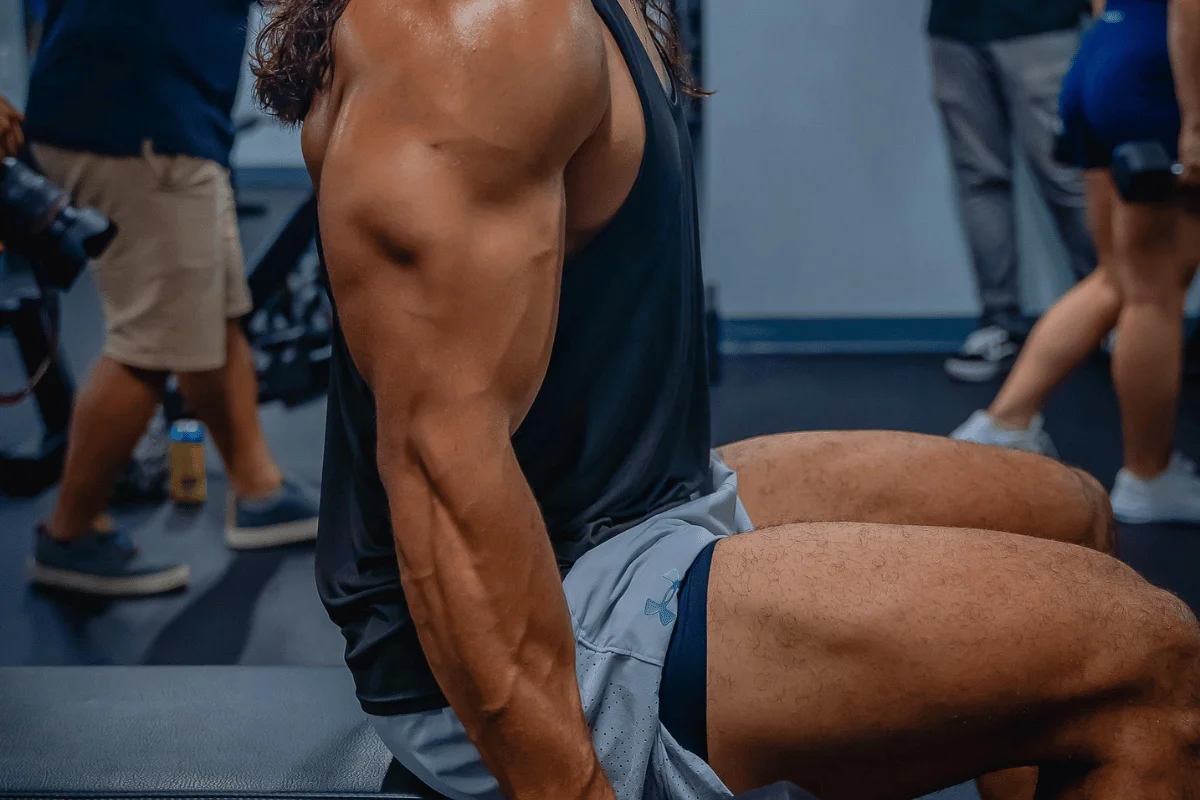 Close-up of a muscular man sitting on a gym bench, showing defined arm and leg muscles.