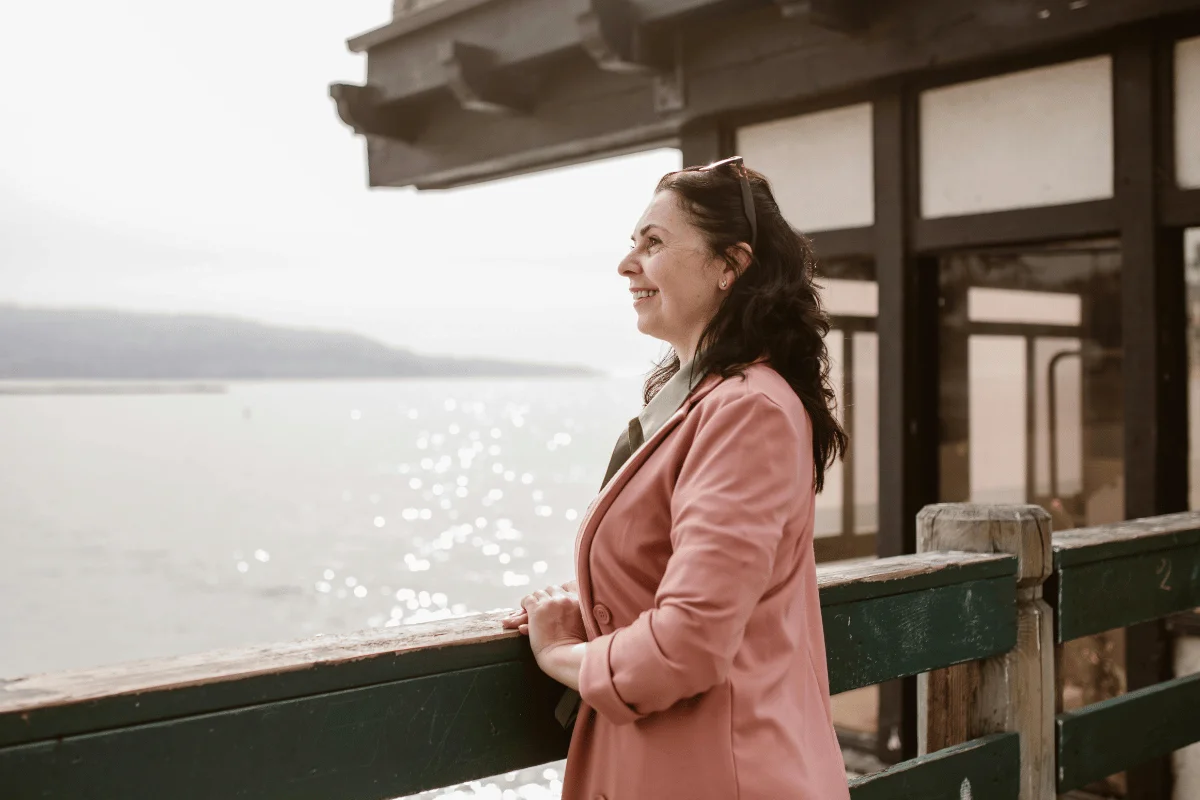 Woman in a pink blazer standing by a railing overlooking a bright glimmering sea.