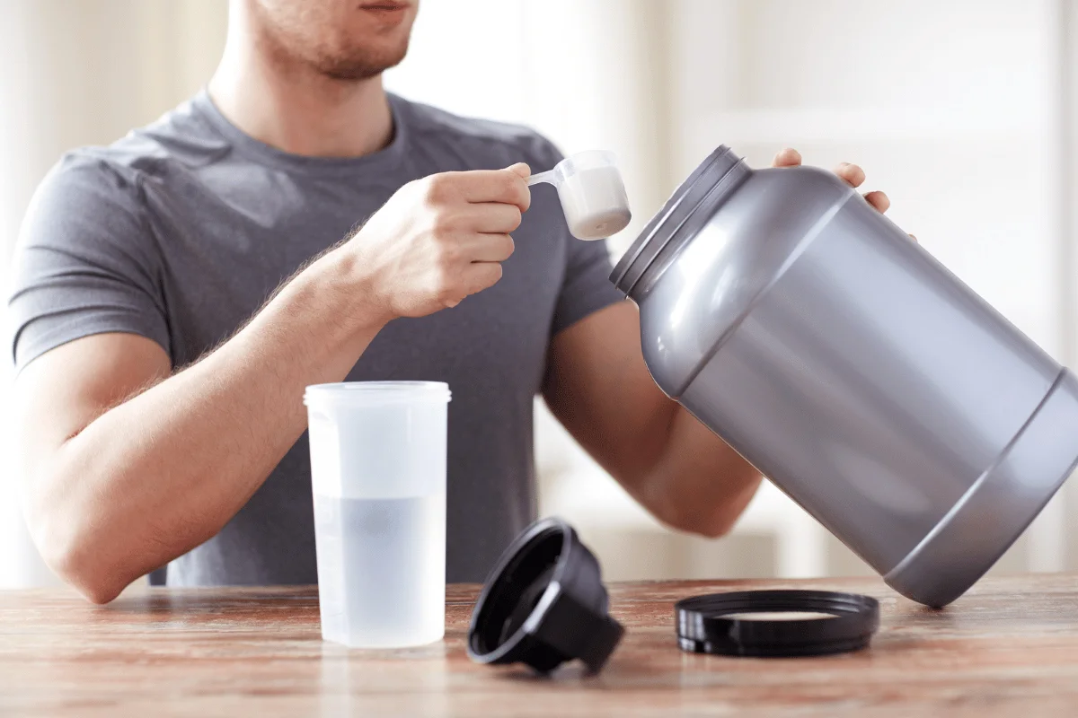 Man scooping protein powder from a large container into a shaker cup with water.