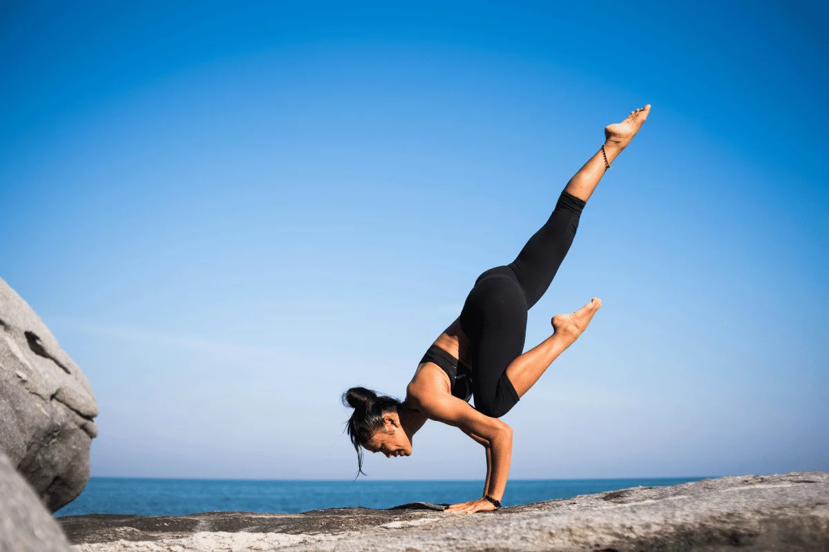 Yoga practitioner holding a handstand on rocks near the sea under a clear sky, demonstrating strength.