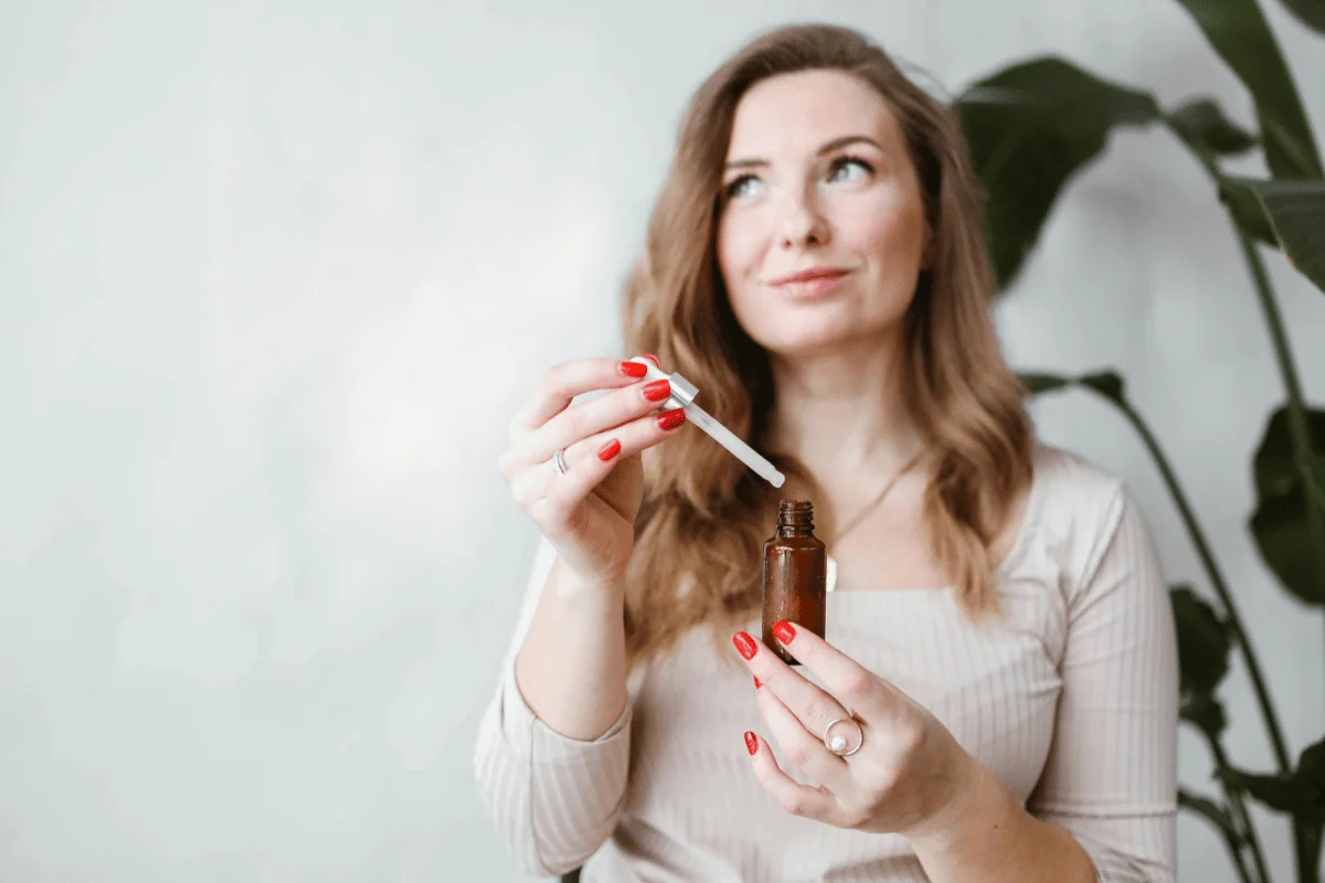 Person holding a dropper bottle, red nails, with indoor plants in background.