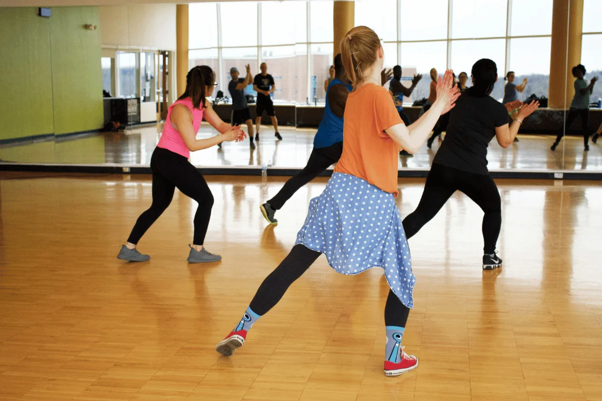 Group of people dancing in a fitness class at a gym with large windows and wooden floors.