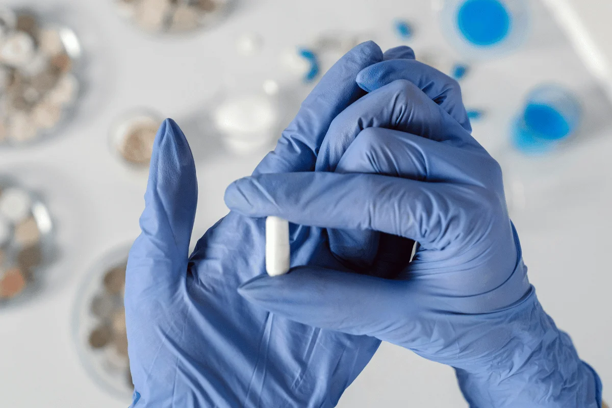 Gloved hands holding a white capsule in a medical lab.