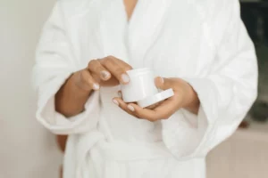 Person in white robe holding an open jar of cream.