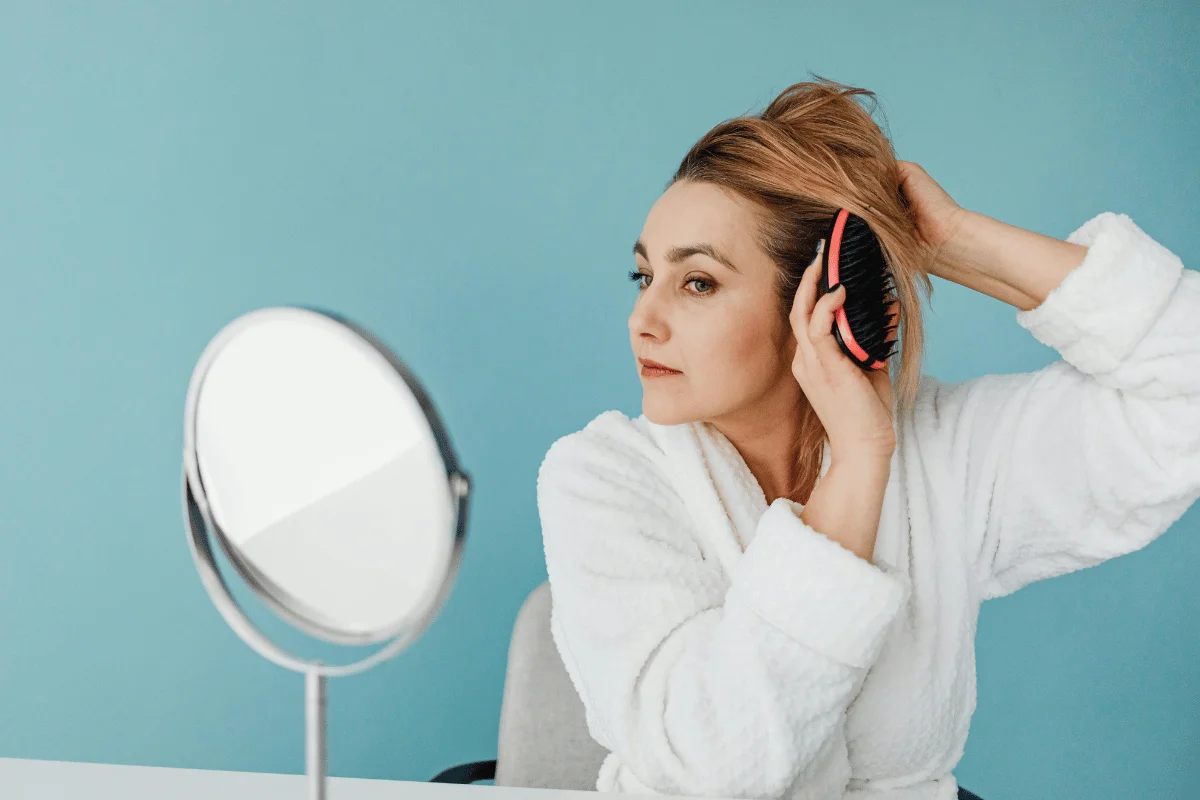 Person in a white robe brushing hair, with a table mirror on a blue background.