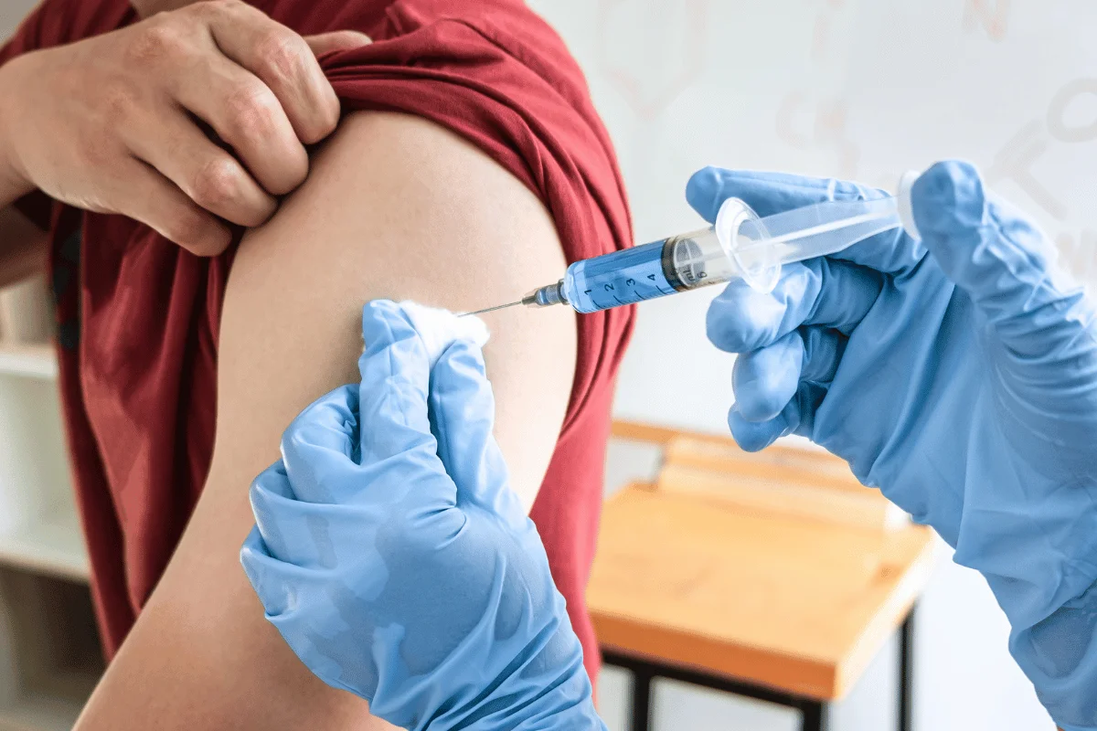 Person receiving a vaccine injection in the upper arm, with a healthcare worker in blue gloves administering it.