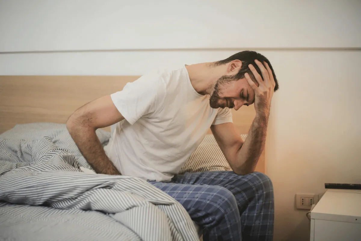 Man sitting on bed, holding head in pain, experiencing discomfort.