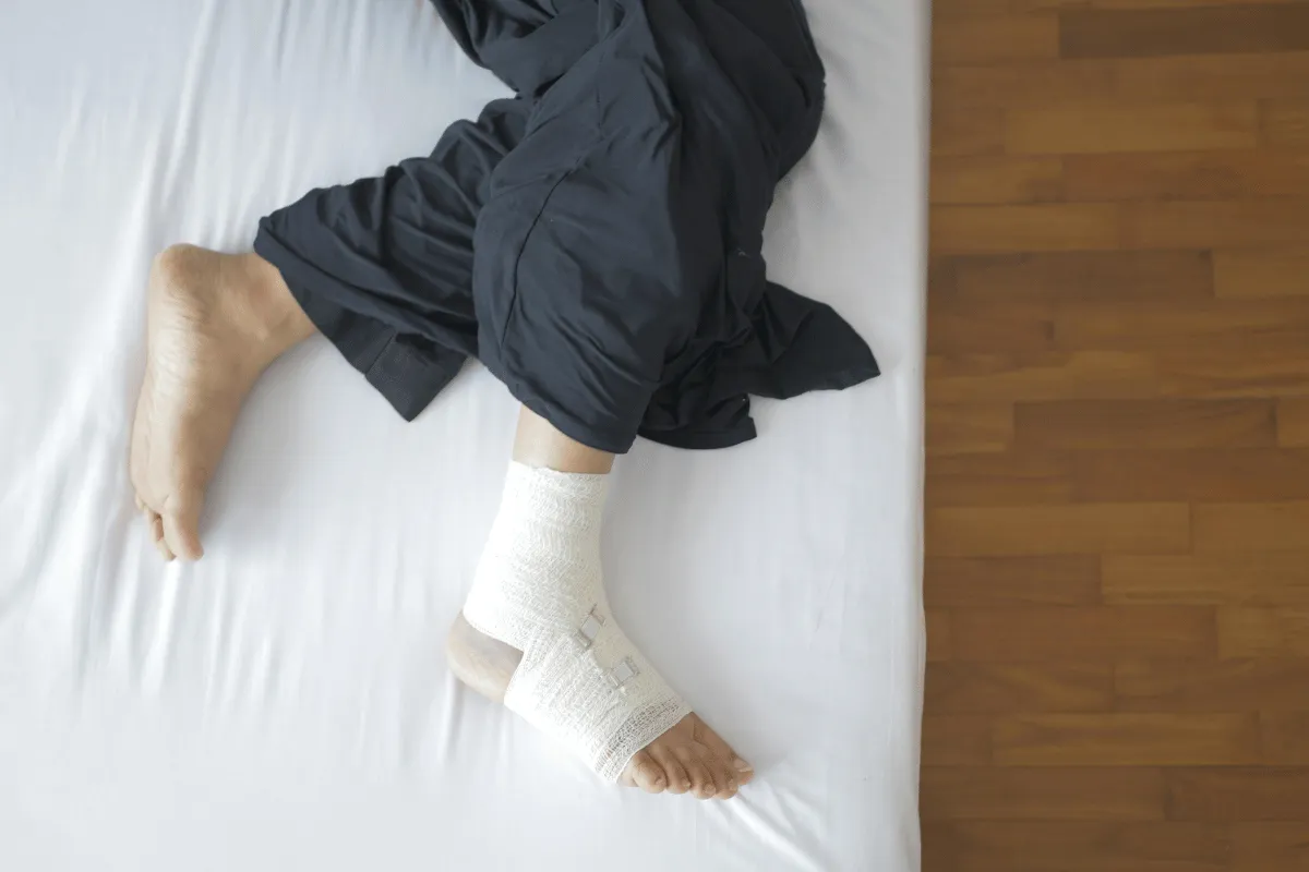 Close-up of a foot with a bandaged ankle, resting on a white bed.