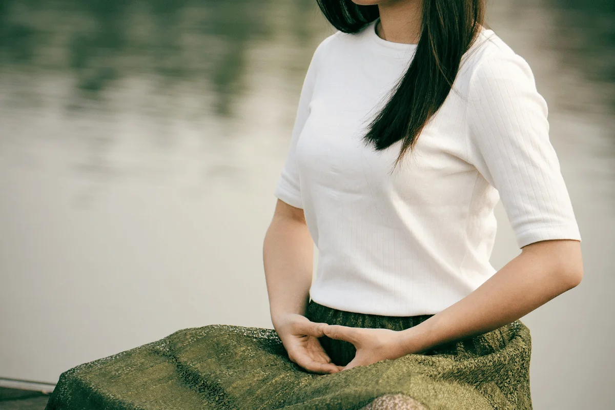 Woman in white shirt and green skirt sitting by water, cropped to show only body from neck down.