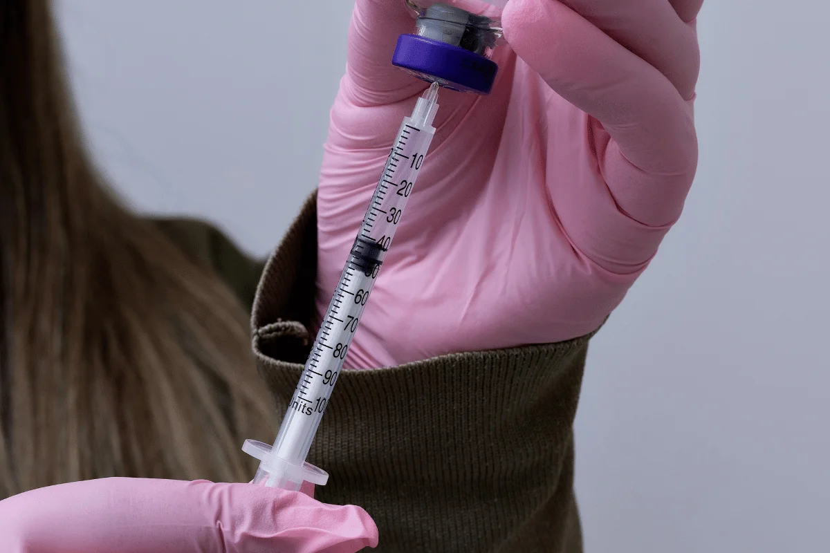 Person in sweater and pink gloves drawing medicine into a syringe from a vial.