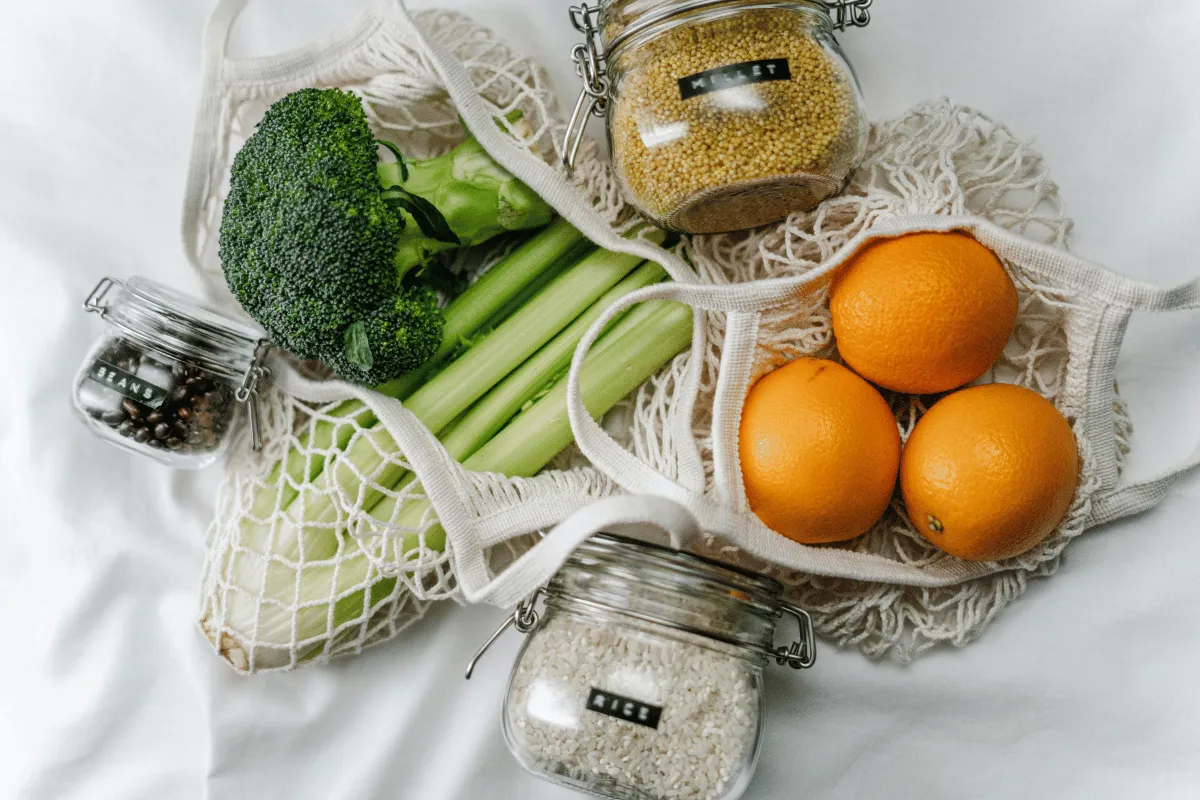 Fresh produce and grains in reusable bags, including broccoli, celery, oranges, and jars of grains.