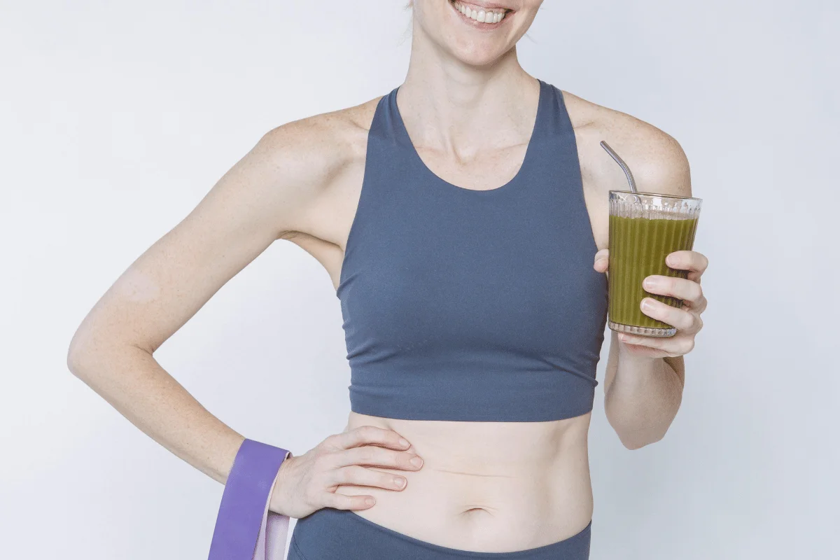 Woman in sportswear holding a green smoothie, smiling, with a yoga mat under her arm.