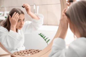 Person in a white robe applying hair treatment looking into a mirror.