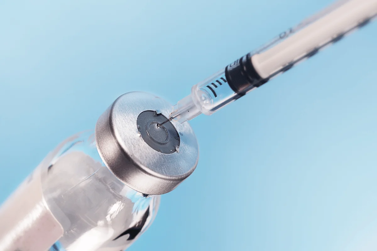 Syringe needle piercing the metal cap of a medicine vial on blue background.