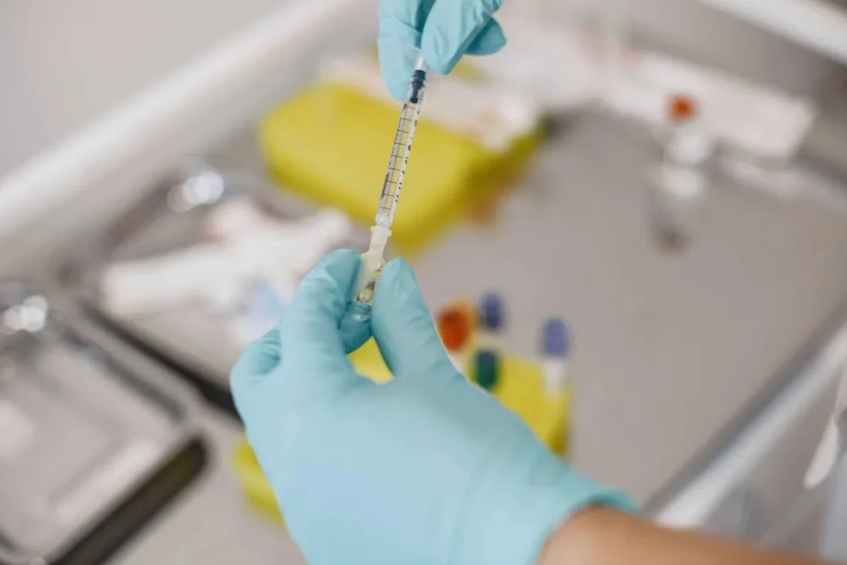 Hands in gloves filling a syringe with medicine from a vial in a clinical setting.