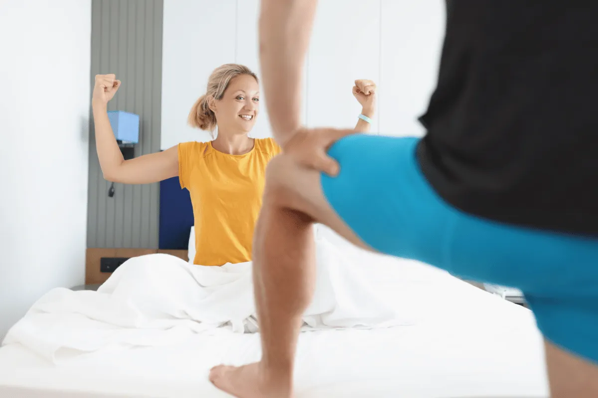 Woman smiling and flexing arms while sitting on bed, with person standing in the foreground.