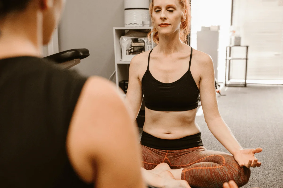 A woman with closed eyes sits cross-legged in a meditative pose during a yoga session indoors.