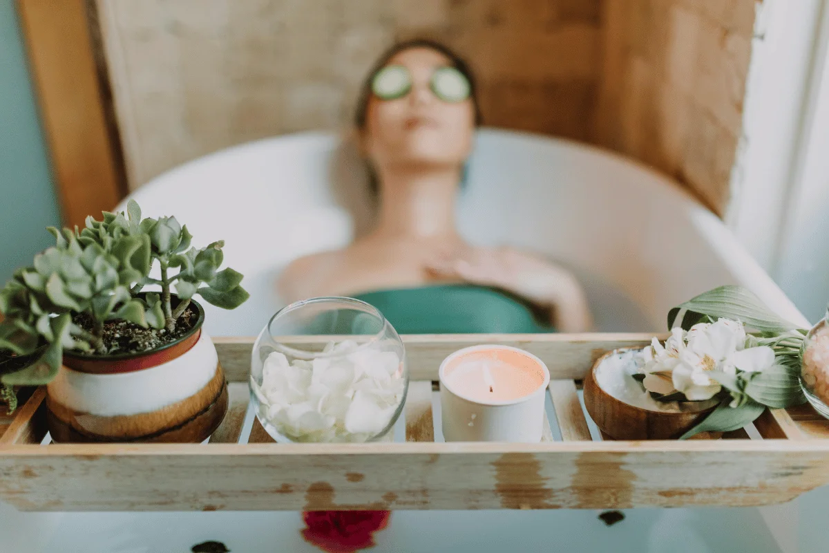 Woman relaxing in a bath with cucumber eye mask, surrounded by candles, plants, and flowers.