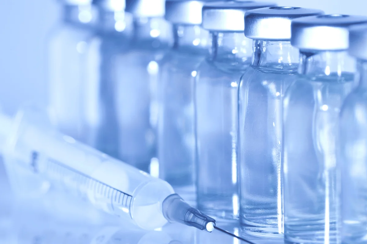 Row of glass medical vials with silver caps next to a syringe.