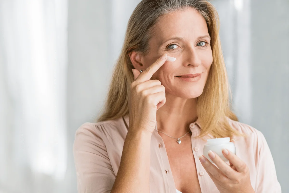 Smiling mature woman applying face cream with her finger, holding a jar of moisturizer.