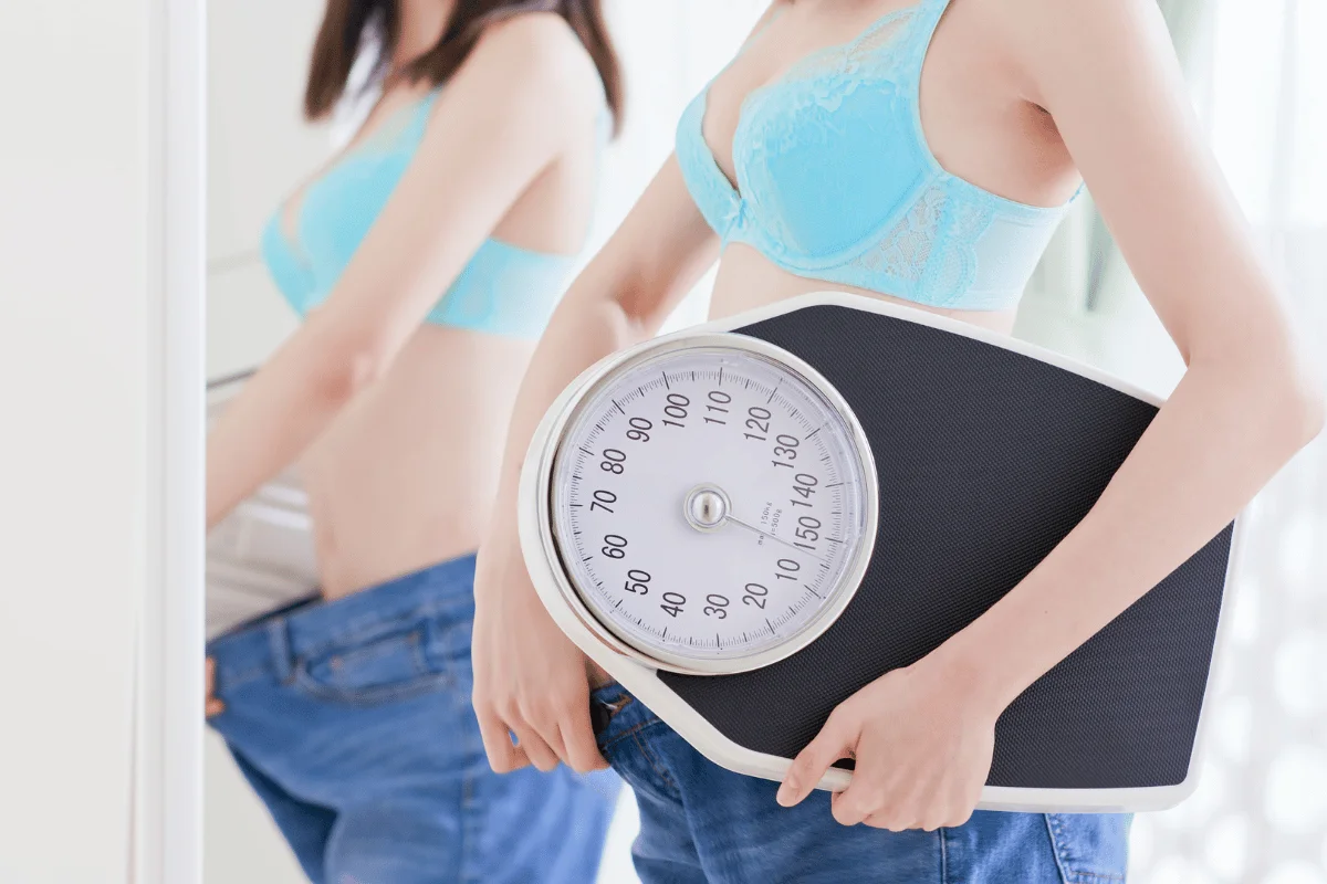 Young woman in a blue bra holding a weight scale while wearing oversized jeans, reflecting her weight loss journey.