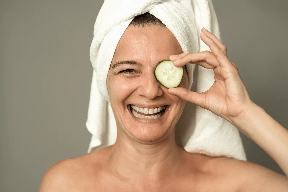 Smiling woman with a towel on her head holding a cucumber slice over her eye.