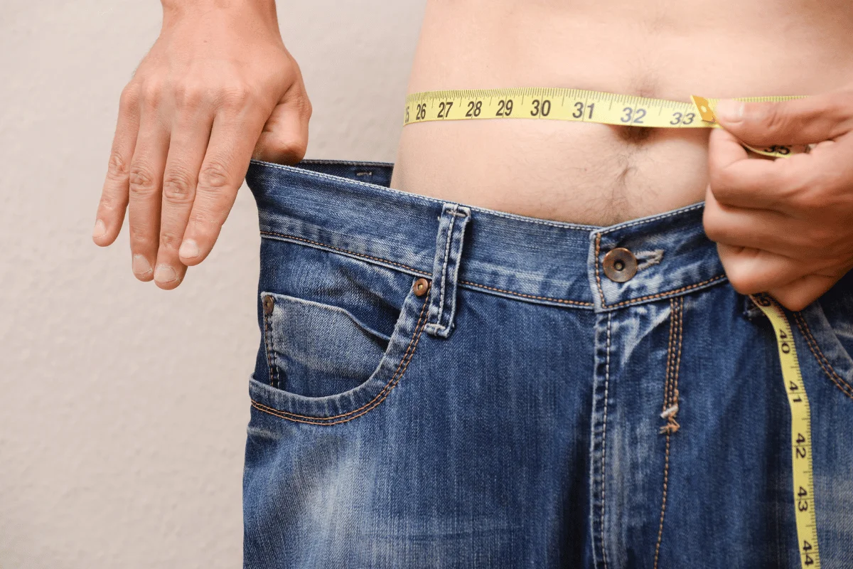 Close-up of a person measuring their waist with a tape while wearing oversized jeans.