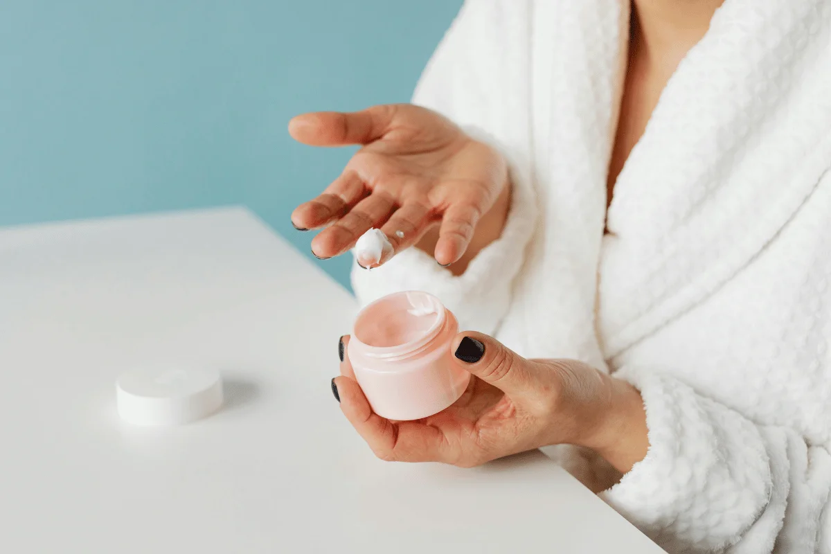 Woman applying moisturizing cream from a pink jar in a spa setting.