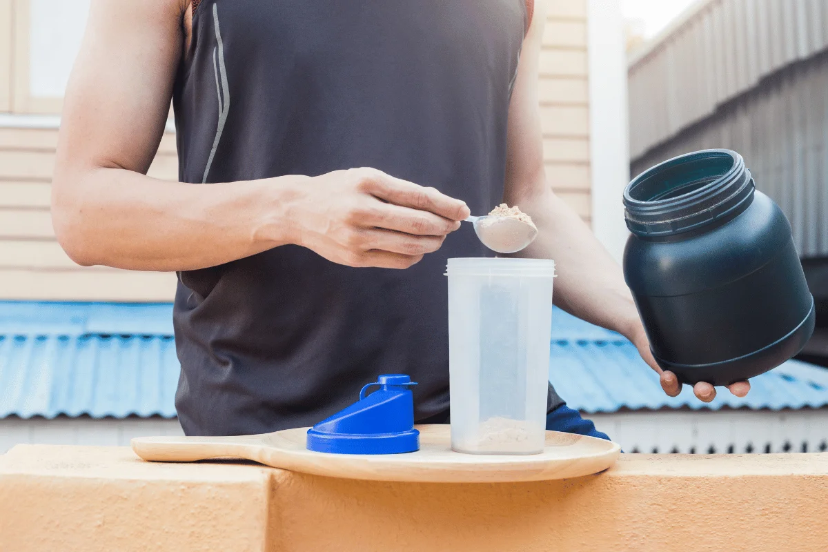 Man preparing a protein shake by scooping whey powder into a shaker bottle outdoors.