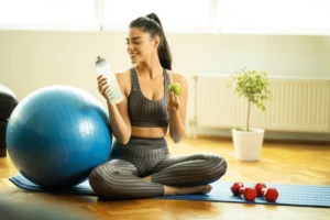 Woman in activewear holding a water bottle and apple, sitting on a yoga mat with fitness ball and dumbbells.