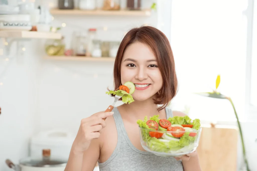 Smiling woman holding a bowl of fresh salad and eating with a fork in a bright kitchen.