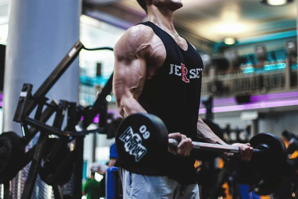 A muscular man in a sleeveless shirt lifts a dumbbell in a gym, showcasing his defined arms and intense focus during a workout.