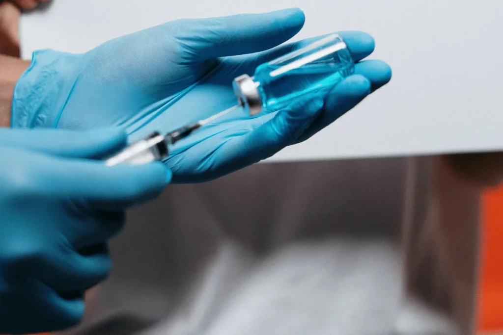 A healthcare professional in blue gloves holds a syringe and a vial of blue liquid, preparing for an injection.