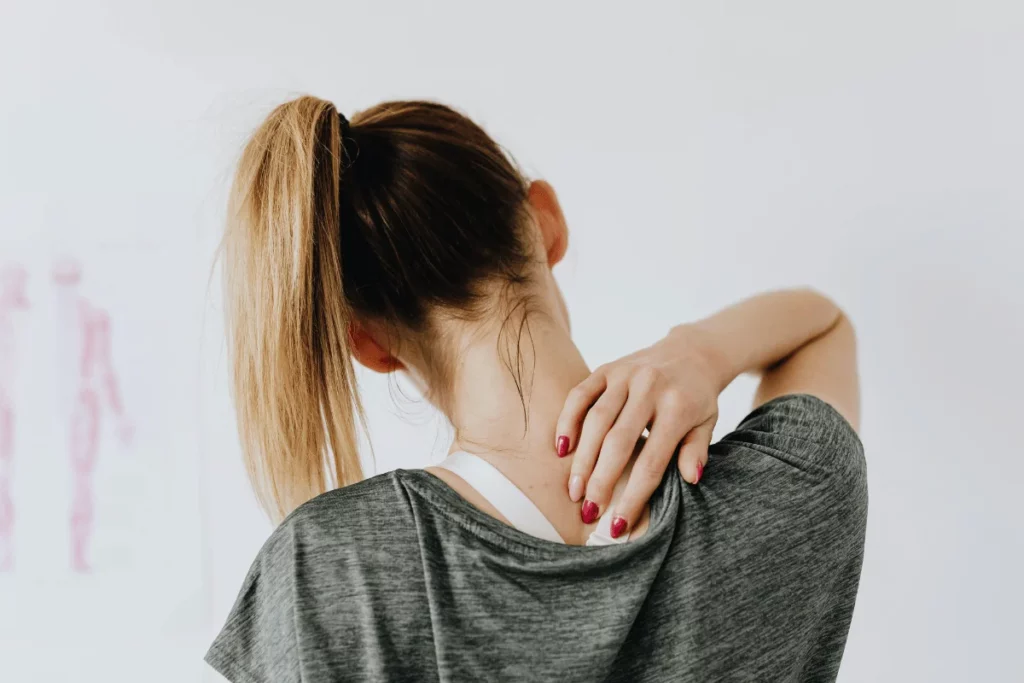 A woman in a gray shirt gently rubs her neck with one hand while facing away, highlighting discomfort or tension.