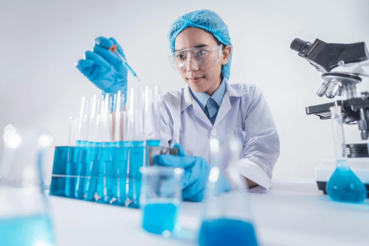 A lab technician in a white coat and blue gloves conducts an experiment with test tubes filled with bright blue liquid.