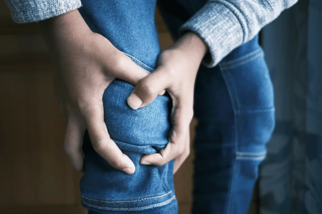 A person holds their knee, showing signs of discomfort, wearing blue jeans and a gray sweater in a softly lit indoor setting.