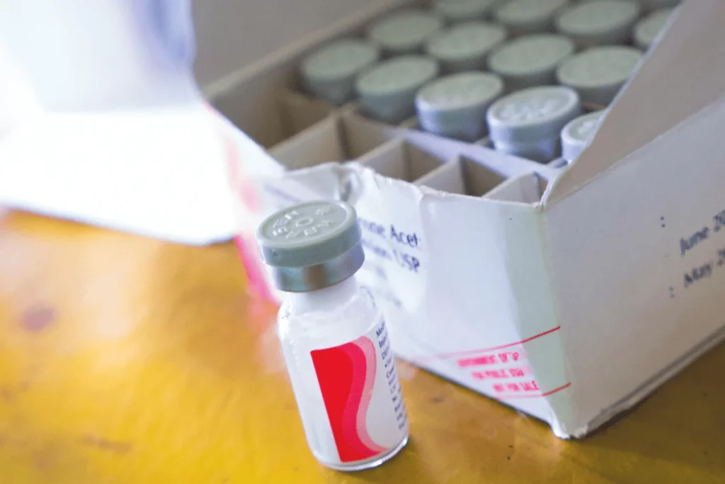 A close-up of a vaccine vial beside an opened box containing multiple vials, on a wooden surface.