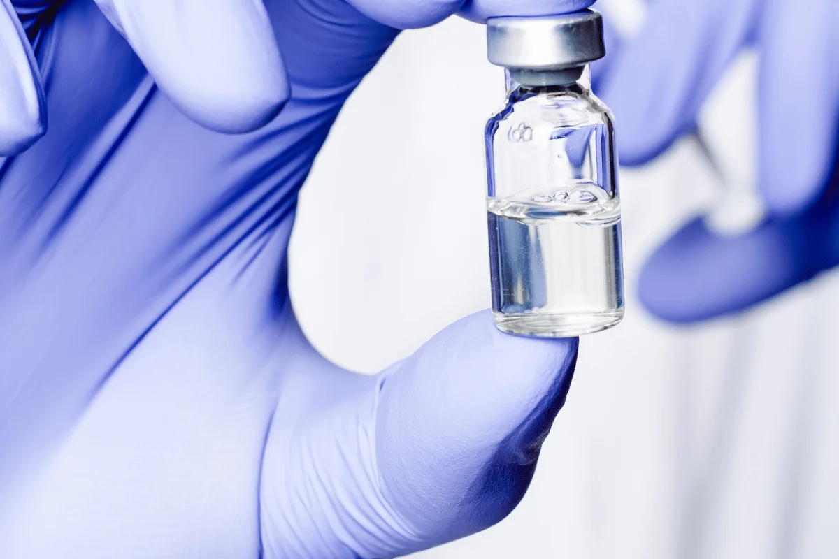 Medical glass vial with liquid, secured by a gloved hand.