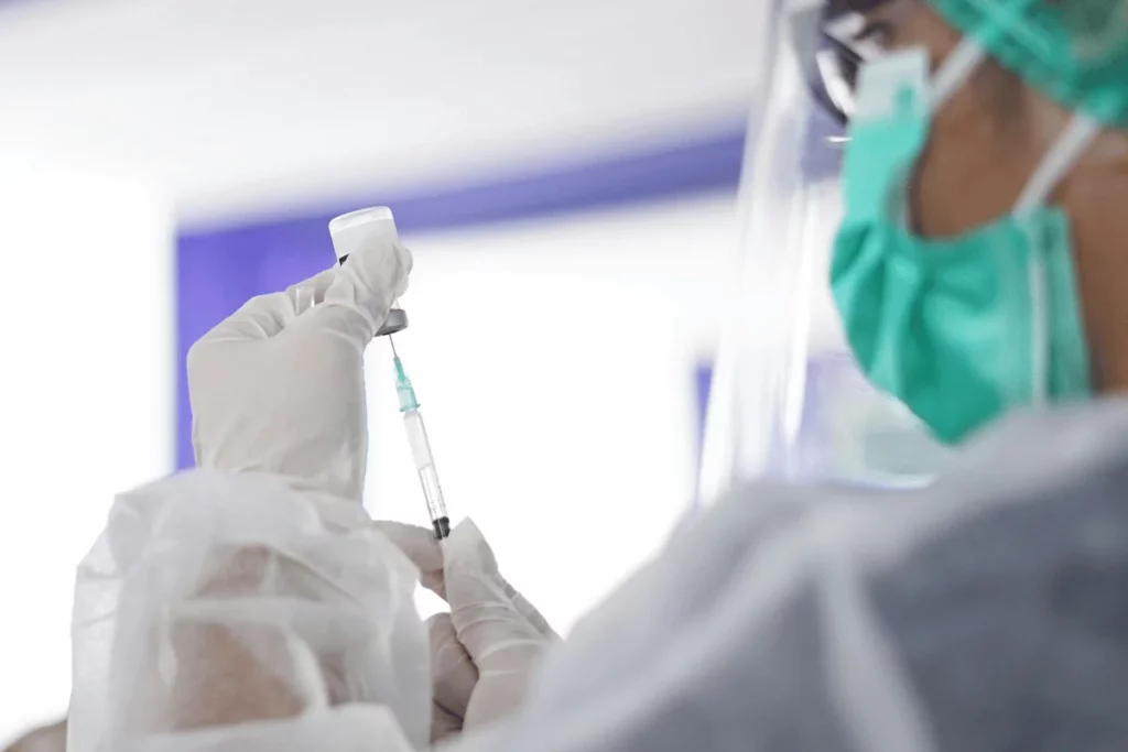 A healthcare worker, wearing gloves and a mask, prepares a syringe with a vaccine from a vial in a clinical setting.