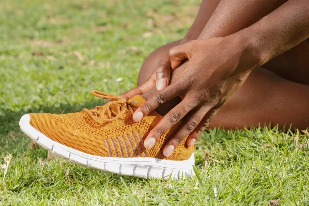 A person in a brown sneaker sits on grass, holding their ankle, indicating discomfort or injury. Their hands show concern.