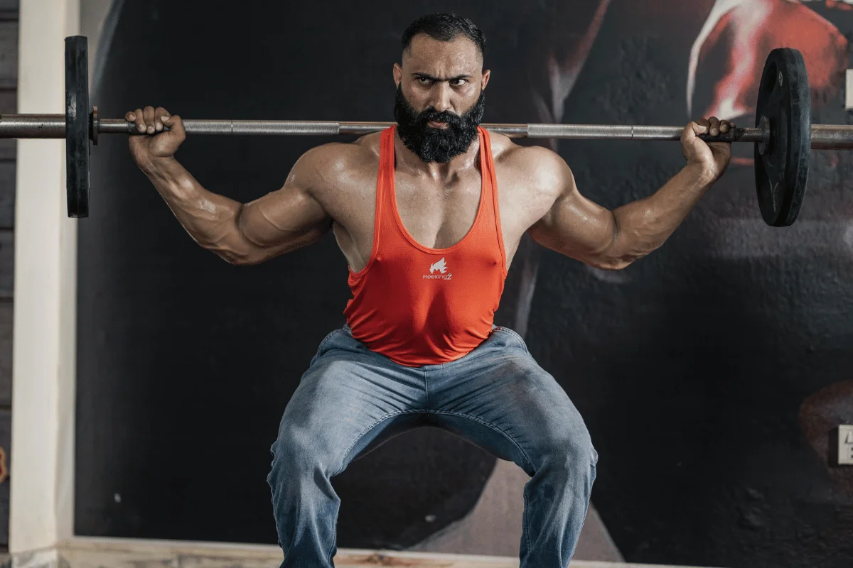 A muscular individual performs a barbell squat in a gym setting, wearing a bright red tank top and jeans.