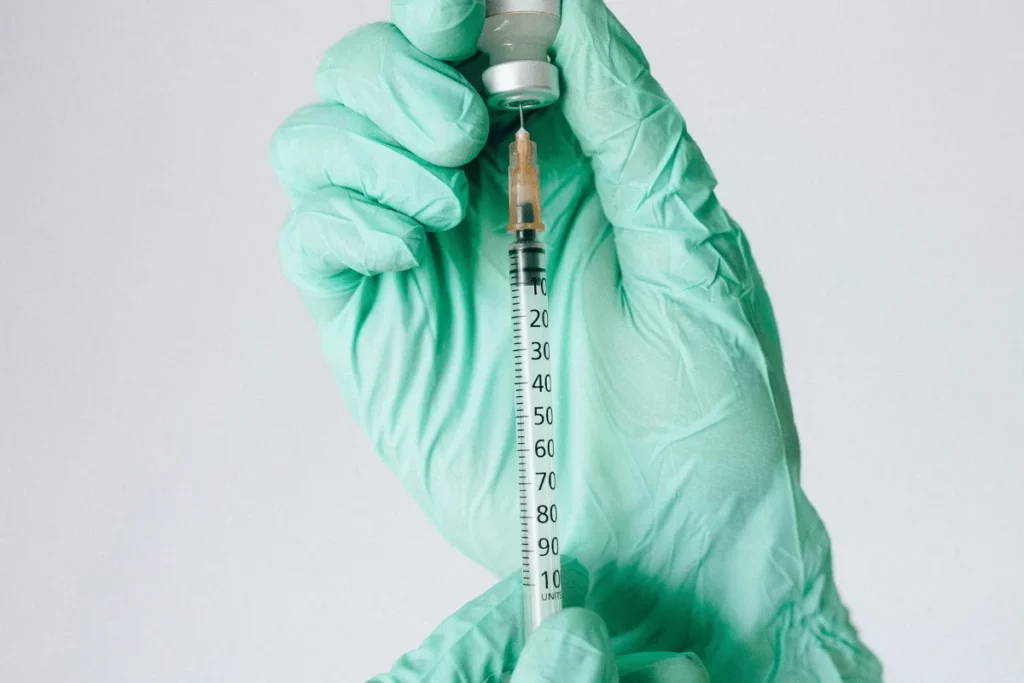 A gloved hand holds a syringe and vial, preparing for an injection. The syringe shows measurement markings up to 100 units.