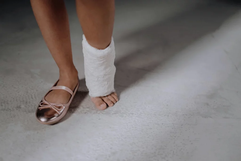 A close-up of a bare foot beside a pink ballet flat and a wrapped ankle, resting on a textured gray surface.