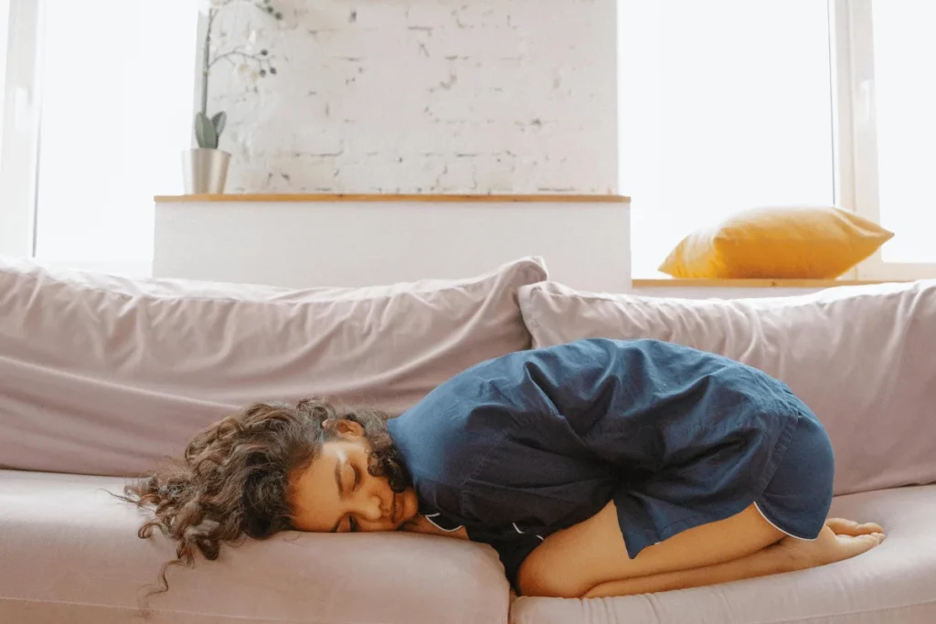 A person in a blue shirt curls up on a soft couch, resting their head against the cushion in a cozy, peaceful setting.