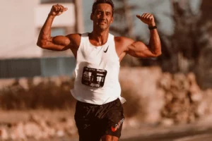 An athletic runner celebrates with raised arms, showcasing muscular physique while wearing a white tank top and black shorts with a race number.